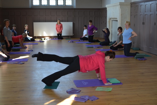 A mat class in a panelled hall, Carole demonstrating a kneeling leg extension